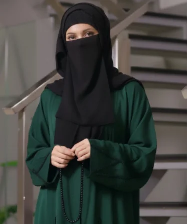 Woman wearing a black niqab and green abaya, holding prayer beads, standing near a staircase.